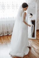 un' donna nel un' bianca nozze vestito sta nel davanti di un' specchio, regolazione sua velo foto