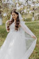 ritratto di un' molto bellissimo sposa con lungo Riccio capelli e indossare un' bianca vestito e lungo velo nel un' fioritura primavera giardino. naarechena detiene sua velo con sua mani su un' verde sfondo di erba foto