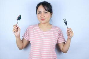 sorridente espressione di giovane asiatico donna guardare a il telecamera mentre Tenere un' forchetta e cucchiaio foto