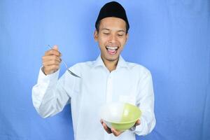 musulmano asiatico uomo indossare berretto In piedi e hold un vuoto ciotola e cucchiaio mostrando il piatto foto