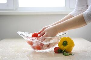 femmina mani avvicinamento lavare ciliegia pomodori nel pulito primavera acqua nel un' bicchiere ciotola giallo Pepe coriandolo le foglie nelle vicinanze pulizia salutare gustoso cibo insalata verdure dieta salutare cibo vitamine foto