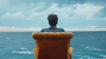 un' uomo seduta solo su un' felpa sedia viso trasformato lontano come sguardi su in il vasto distesa di il mare perso nel pensato e ispirazione. . foto