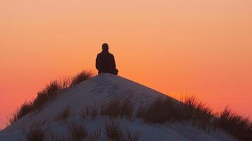 un' solitario figura si siede in cima un' grande sabbia duna silhouette delineato contro il arancia e rosa tonalità di il ambientazione sole. con . foto