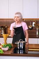 donna allegra senior sta bevendo vino rosso durante la cottura in cucina moderna. cibo, educazione, concetto di stile di vita foto
