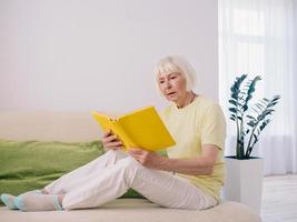 donna anziana con i capelli grigi che legge un libro su un divano a casa. educazione, pensione, anti età, concetto di lettura