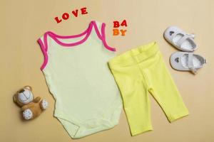 body bianco per bambina su fondo beige con scarpe da bambino, orsacchiotto, pantaloni gialli, modello di body per neonati. piatto, vista dall'alto. foto