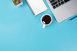 tavolo da ufficio con spazio di lavoro, computer portatile, tazza di caffè, taccuino, matita, pianta e telefono cellulare su sfondo blu pastello