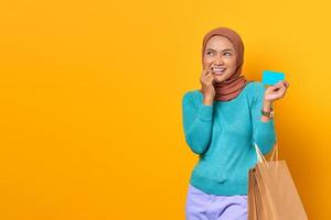 sorridente giovane donna asiatica shopping tenendo la carta di credito con un dito sulle labbra su sfondo giallo