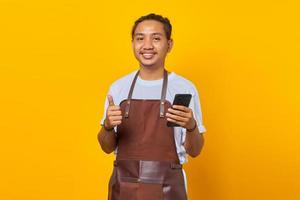 uomo asiatico eccitato e allegro che tiene smartphone e pollice in alto su sfondo giallo