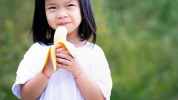 bambino felice tiene in mano una banana matura da mangiare con gusto. lo sfondo è verde naturale. spazio vuoto per inserire il testo. foto