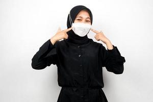 donna musulmana che indossa una maschera bianca, con la mano che punta alla maschera, previene il gesto del virus corona, previene il covid-19, isolato su sfondo bianco