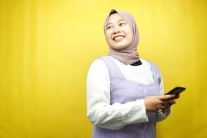 bella giovane donna musulmana asiatica sorridente sicura di sé, entusiasta e allegra con le mani che tengono lo smartphone, vista guardando lo spazio vuoto che presenta qualcosa, isolato su sfondo giallo