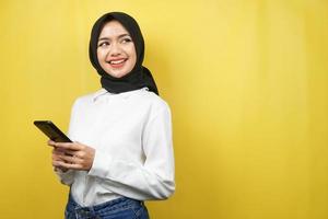 bella giovane donna musulmana asiatica sorridente sicura di sé, entusiasta e allegra con le mani che tengono lo smartphone, vista guardando lo spazio vuoto che presenta qualcosa, isolato su sfondo giallo