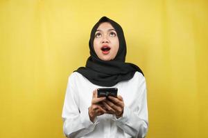 bella giovane donna musulmana asiatica scioccata, sorpresa, espressione wow, mano che tiene smartphone, alzando lo sguardo, isolata su sfondo giallo foto
