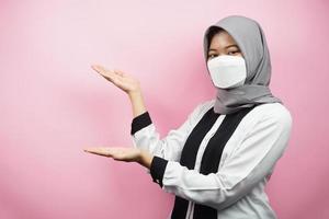 donna musulmana che indossa una maschera bianca, con la mano che punta allo spazio vuoto che presenta qualcosa, isolato su sfondo rosa foto