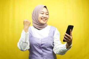 bella giovane donna musulmana asiatica sorridente sicura di sé, entusiasta e allegra con le mani che tengono lo smartphone, le mani serrate, pugni, vittoria, successo, buon lavoro, isolato su sfondo giallo foto