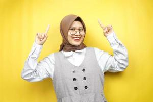 bella giovane donna musulmana asiatica sorridente sicura di sé, entusiasta e allegra con le mani rivolte verso l'alto presentando qualcosa, guardando la telecamera isolata su sfondo giallo, concetto pubblicitario foto