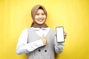 bella giovane donna musulmana asiatica sorridente sicura di sé, entusiasta e allegra con la mano che tiene lo smartphone, promuovendo qualcosa, app per la promozione della mano, isolata su sfondo giallo