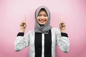 bella giovane donna musulmana asiatica sorridente sicura di sé, entusiasta e allegra con le mani rivolte verso l'alto presentando qualcosa, guardando la telecamera isolata su sfondo rosa, concetto pubblicitario foto
