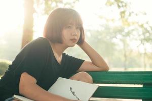 hipster felice giovane donna asiatica che scrive nel suo diario nel parco. hipster felice giovane donna asiatica che lavora al taccuino nel parco. studente che studia all'aperto. concetto all'aperto della donna di stile di vita. bagliore di luce. foto