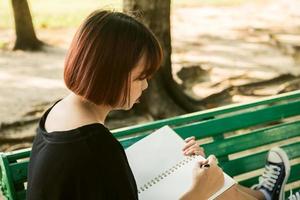 hipster felice giovane donna asiatica che scrive nel suo diario nel parco. hipster felice giovane donna asiatica che lavora al taccuino nel parco. studente che studia all'aperto. concetto all'aperto della donna di stile di vita. bagliore di luce. foto