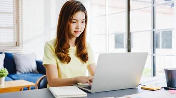 donna d'affari asiatica che utilizza laptop parla con i colleghi del piano in videochiamata mentre lavora in modo intelligente da casa in soggiorno. autoisolamento, distanziamento sociale, quarantena per la prevenzione del virus corona. foto