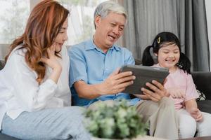 nonni asiatici e nipote utilizzando tablet a casa. cinese anziano, nonno e nonna felici trascorrono il tempo in famiglia rilassandosi con la ragazza che controlla i social media, sdraiati sul divano nel concetto del soggiorno foto