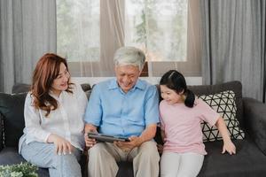 nonni asiatici e nipote utilizzando tablet a casa. cinese anziano, nonno e nonna felici trascorrono il tempo in famiglia rilassandosi con la ragazza che controlla i social media, sdraiati sul divano nel concetto del soggiorno foto
