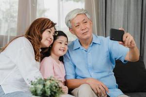 selfie dei nonni asiatici con la nipote a casa. cinese anziano, nonno e nonna felici trascorrono il tempo in famiglia rilassarsi utilizzando il telefono cellulare con una ragazzina sdraiata sul divano nel concetto di soggiorno. foto
