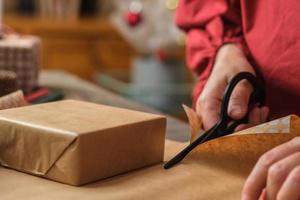 primo piano donna che taglia a mano la carta kraft per avvolgere la confezione regalo di natale su un tavolo di legno foto