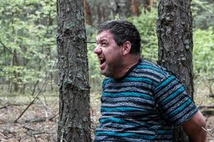 un ostaggio nel bosco. un uomo in maglietta blu e pantaloni è seduto per terra legato a un albero nella foresta e chiede aiuto. vittima di un attacco. foto orizzontale.