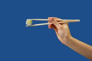 Mano della donna che tiene il rotolo di sushi di maki vegetariano fresco con le bacchette di legno, isolato su fondo blu. colore pantone dell'anno 2020 foto