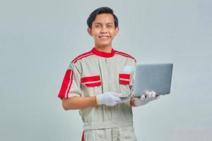 Sorridente bel giovane meccanico che indossa l'uniforme tenendo il laptop e guardando la telecamera isolata su sfondo grigio foto