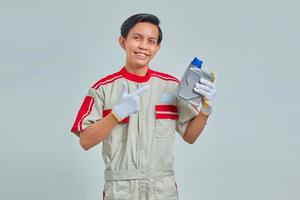 Ritratto di un bell'uomo sorridente che indossa l'uniforme del meccanico che mostra la bottiglia di plastica dell'olio motore su sfondo grigio foto