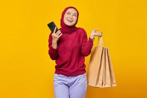 sorridente giovane donna asiatica che tiene smartphone e borsa della spesa mentre guarda in alto su sfondo giallo foto