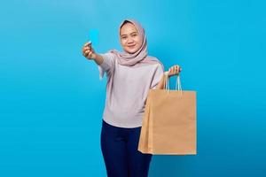 ritratto di allegra donna asiatica che tiene in mano la borsa della spesa e mostra la carta di credito su sfondo blu foto