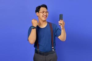 eccitato giovane asiatico che utilizza lo smartphone per celebrare il successo isolato su sfondo viola foto