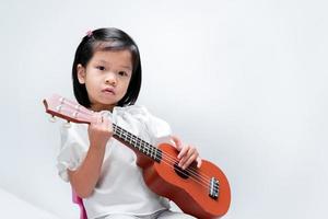 il bambino carino impara a suonare la piccola chitarra nella scuola di musica. la ragazza sta con gli ukulele su sfondo grigio. foto