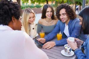 ragazzo che mostra il suo smartphone al suo gruppo di amici mentre beve un drink in un bar all'aperto foto