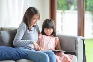 felice madre asiatica che insegna a sua figlia carina a studiare nel soggiorno di casa foto