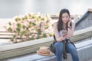 bella donna asiatica turistica solista che si rilassa e si diverte ad ascoltare la musica su uno smartphone nel centro urbano della città. viaggio di vacanza in estate. foto