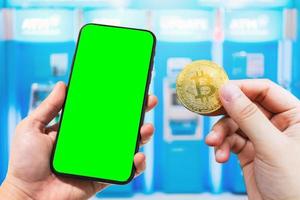 usa la mano che tiene lo smartphone e la moneta bitcoin immagini sfocate tocco di sfocatura astratta in sfocato di bancomat blu nel grande magazzino, concetto di criptovaluta di pagamenti elettronici. foto