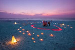 bellissimo tavolo allestito per una cena romantica sulla spiaggia con lanterne e sedie e fiori con candele e cielo e mare sullo sfondo. cena in spiaggia al tramonto