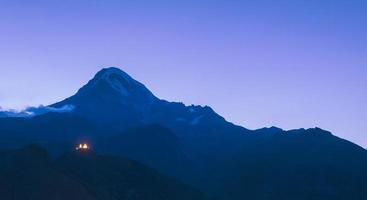 bellissimo panorama blu della chiesa della trinità di gergeti con vista mozzafiato sul picco kazbek sullo sfondo foto