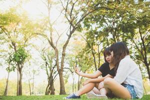 due belle giovani amiche asiatiche felici che si divertono insieme al parco e si fanno un selfie. felice hipster giovani ragazze asiatiche sorridenti e guardando smartphone. concetti di stile di vita e amicizia. foto