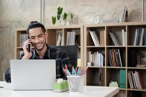 giovane lavoratore di sesso maschile di etnia asiatica, laptop per svolgere lavori creativi, comunicazione con il telefono cellulare sulla scrivania, libreria dietro al posto di lavoro casuale, imprenditore di avvio e occupazione di e-commerce online. foto