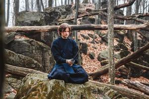 l'uomo in kimono nero con una spada medita e si concentra su rocce e sullo sfondo della foresta foto