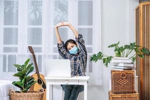 una donna che indossa una maschera usa un laptop al lavoro e allunga le braccia per rilassarsi.