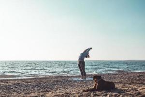 giovane donna che fa yoga e si allunga sulla spiaggia con il suo cane foto