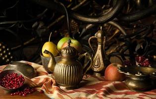 frutta e una brocca di metallo con ciotole in stile orientale sullo sfondo di un ornamento decorativo di forgiatura. natura morta in stile orientale foto
